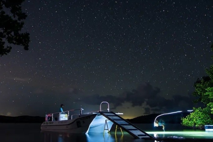 Starry night sky over a lake with a dock and lit boat.